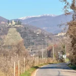 Veduta della Santissima di Gussago dal basso con vigneti in primo piano e montagne innevate sullo sfondo.
